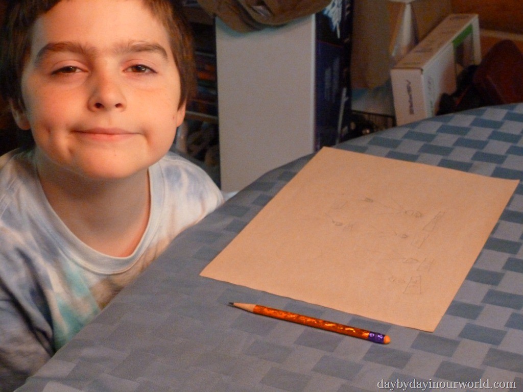 Boy smirks at camera while sitting at a table with a finished drawing on it.
