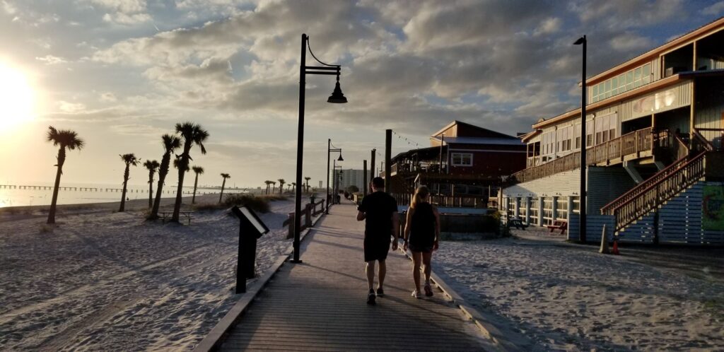 Couple walks on boardwalk at beach. Setting sun on left with palm trees and sand. Buildings on right.