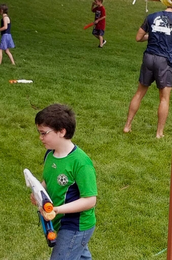 Boy in green shirt holds water gun with multiple kids running behind him.