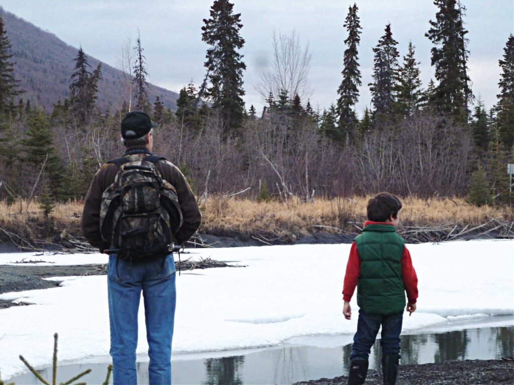 Young boy and his dad observe the change from winter to spring at river mostly covered with snow.