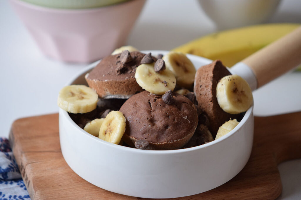 Bowl of chocolate pancake poppers topped with banana slices and chocolate chips on a wooden board, with bananas and a small bowl in the background.