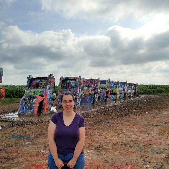 Woman wearing a purple top and holding a purse with a row of painted old Cadillacs stuck in the ground behind her.