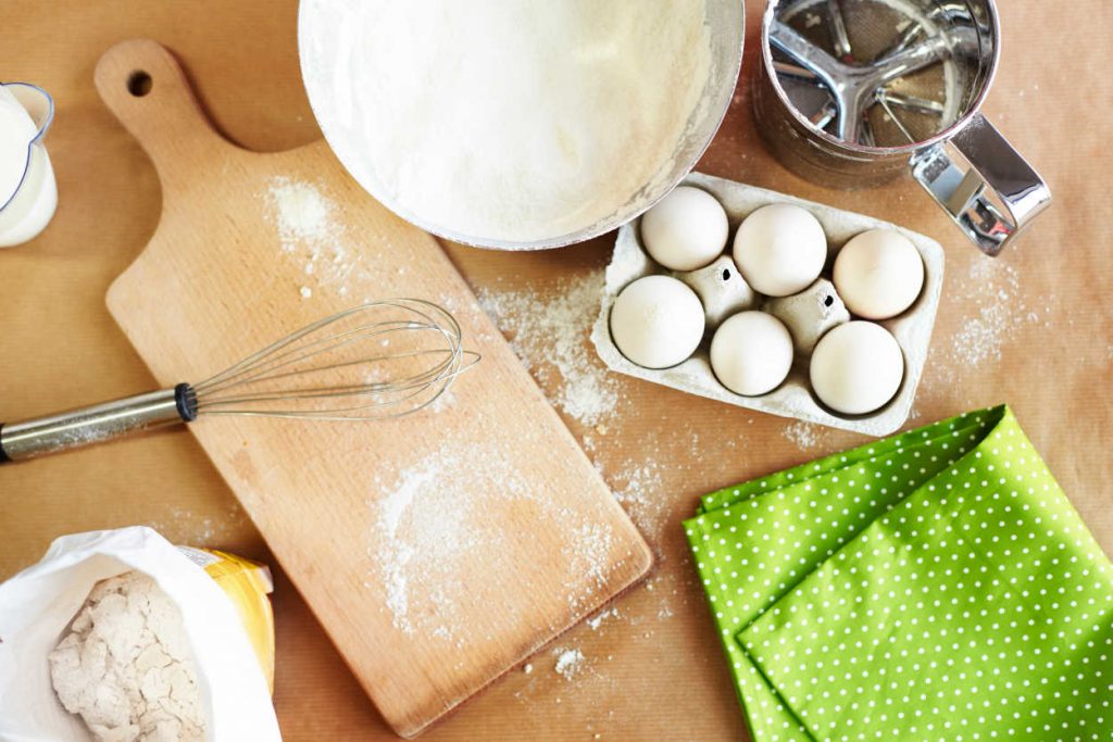 Different baking ingredients (flour, eggs, milk) and tools (whisk, cutting board, sifter and towel) on counter top.