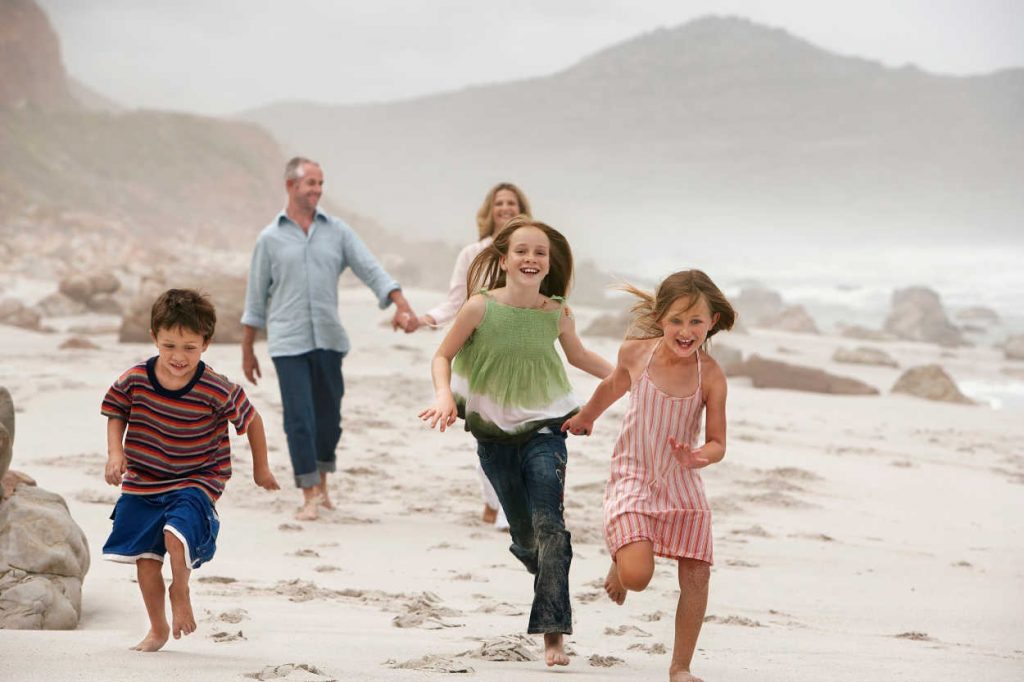 Parents walking while holding hands behind 3 kids who are running along the beach.