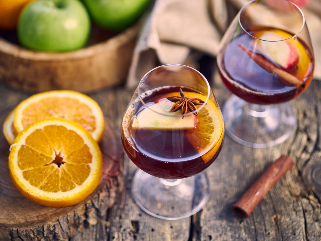 2 glasses of hot apple punch with fruit slices on top. Slices of orange sit to the side and a bowl of apples is in the background.