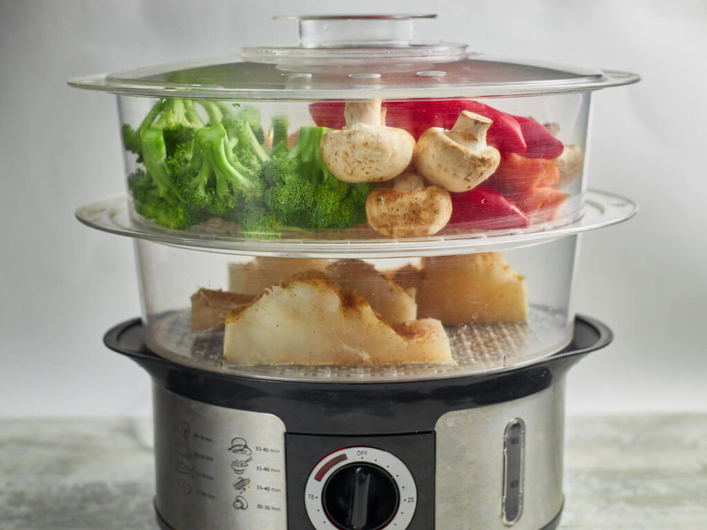 Electrick two-tier steamer with fish chunks on bottom tier and large chunks of broccoli, carrots, mushrooms, and red peppers on top tier.