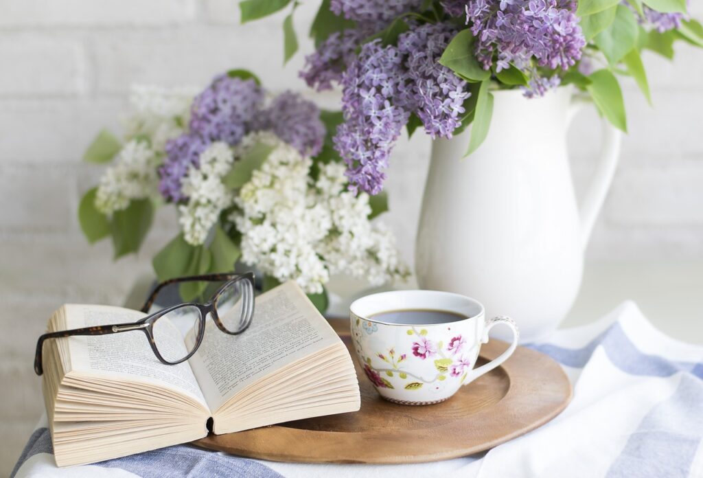 open book with glasses on it. cup of tea sits to the side with a pitcher holding purple flowers.