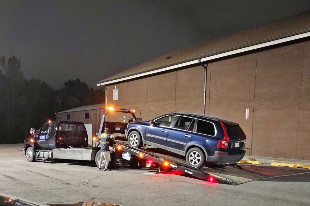 A tow truck is parked in front of a building at night with a Volvo wagon being loaded.