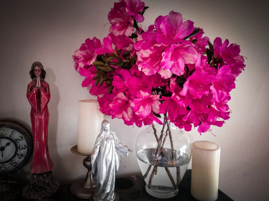 Vase holds azalea flowers with statues of an angel and Mary near by.