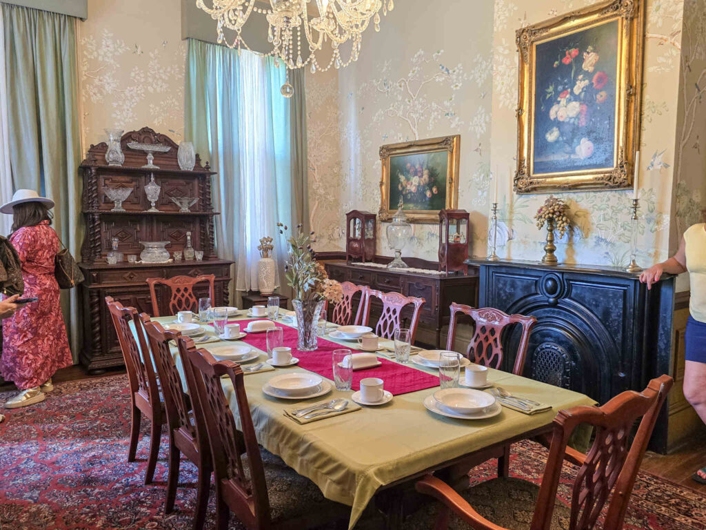 Formal dining room at Poche Plantation with a long table set for guests, antique furnishings, crystal chandelier, floral wallpaper, and historic artwork.