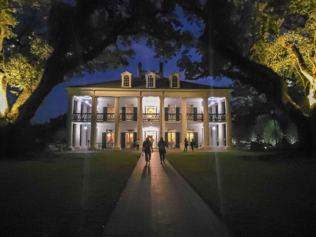 Oak Alley Plantation illuminated at night beneath centuries-old live oak trees draped with Spanish moss, with visitors walking toward the historic mansion.