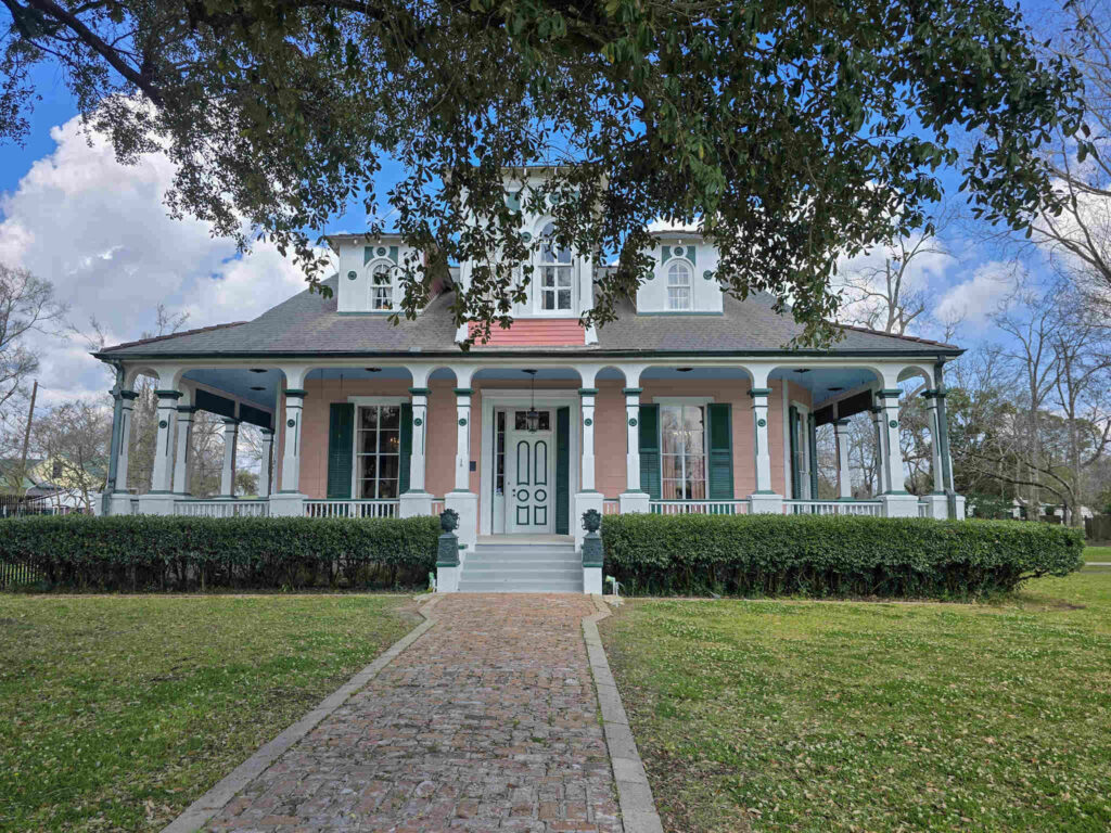 Colorful exterior view of Poche Plantation in Convent, Louisiana, featuring a wraparound porch, decorative trim, and brick walkway leading to the historic home.