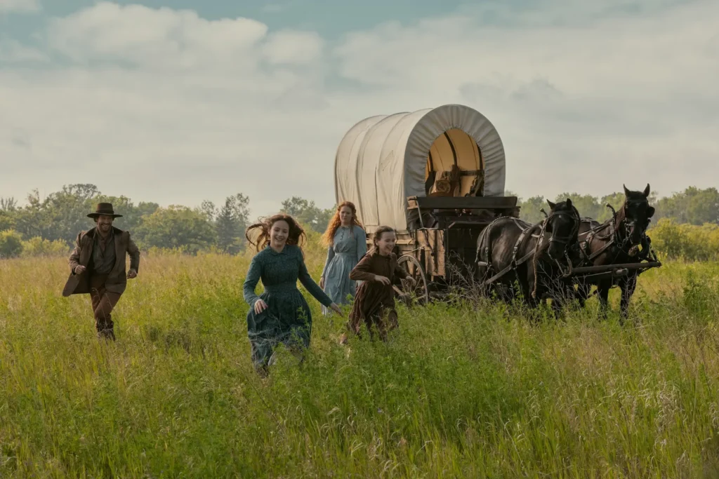 Scene from Netflix’s Little House on the Prairie showing the Ingalls family walking through a prairie field beside a horse-drawn covered wagon.