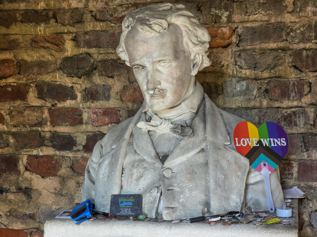 Bust of Edgar Allan Poe displayed against a brick wall at the Poe Museum in Richmond, Virginia, with coins and small items left at its base.