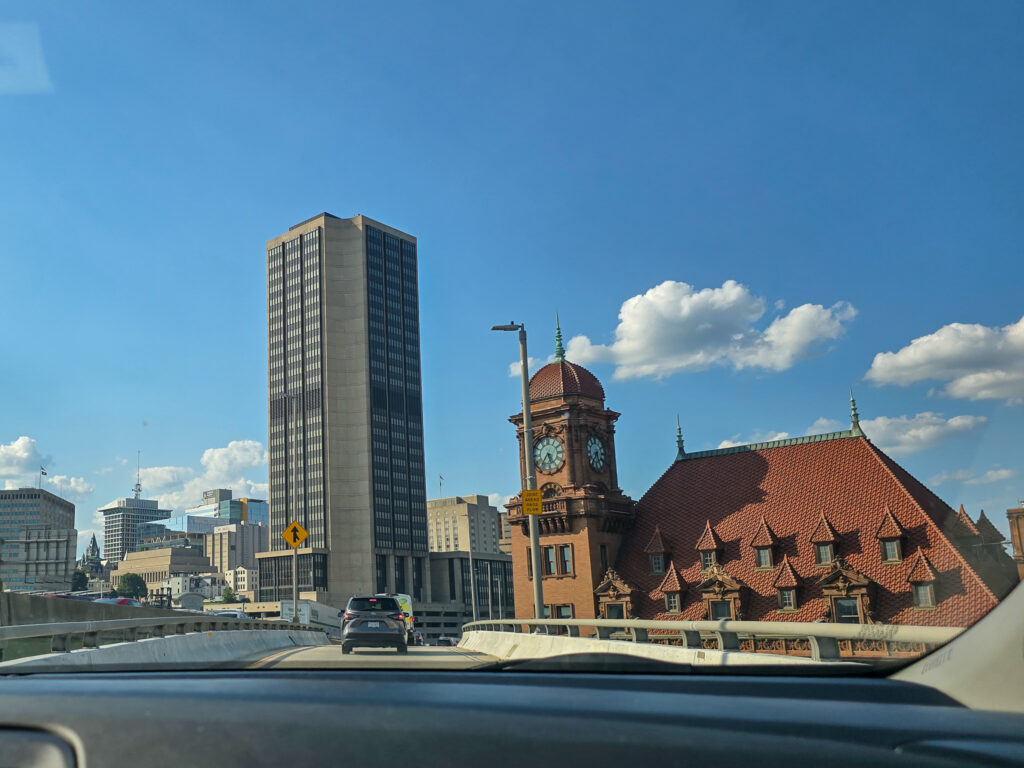 View of downtown Richmond from the road with historic Main Street Station and its clock tower alongside modern high-rise buildings under a bright blue sky.