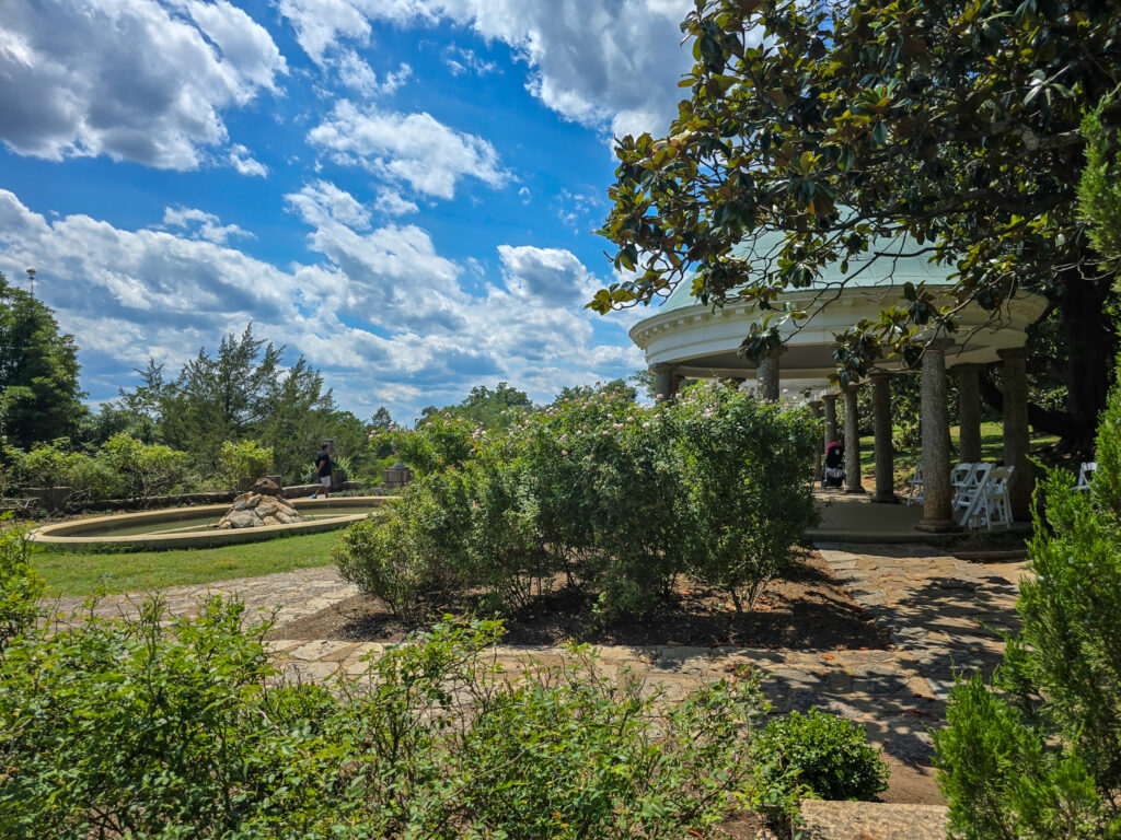 Italian Garden at Maymont in Richmond with manicured greenery, stone paths, a fountain, and a curved columned pavilion beneath a blue, cloud-filled sky.
