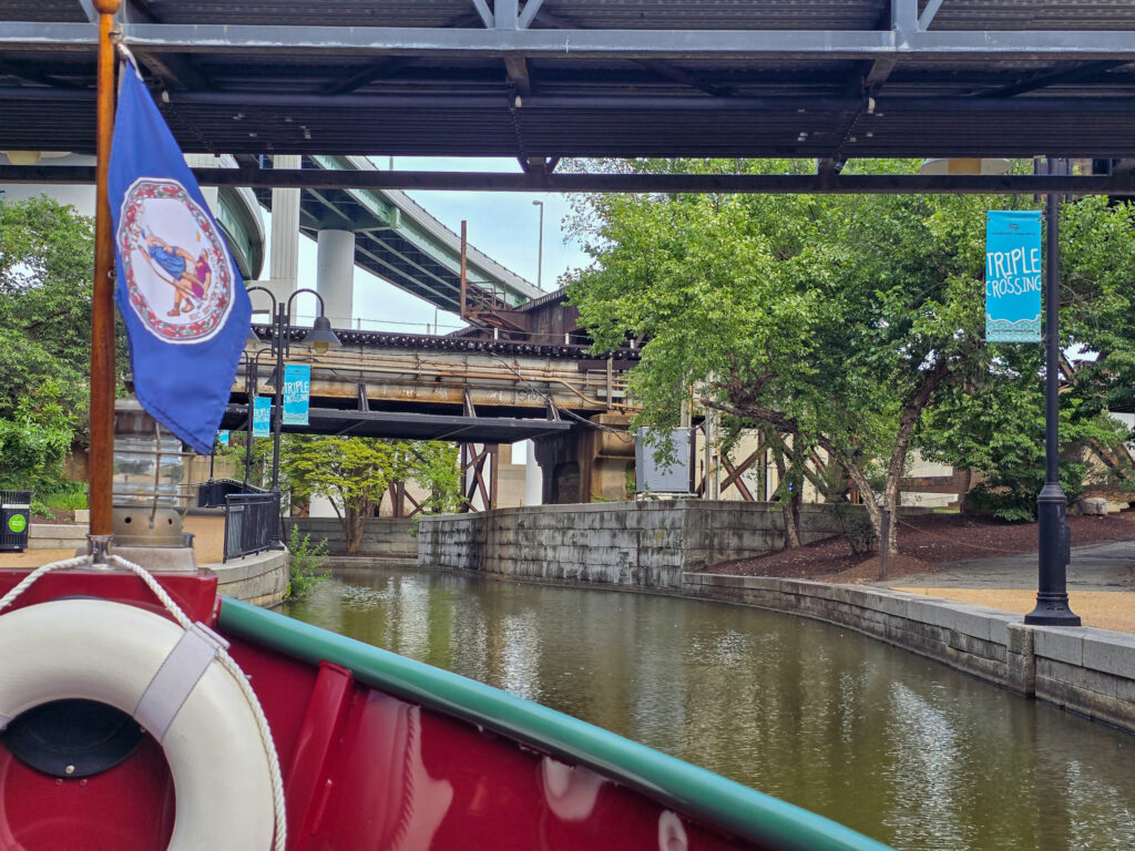View from a canal boat traveling along Richmond’s Canal Walk, with railroad and highway bridges crossing the water ahead.